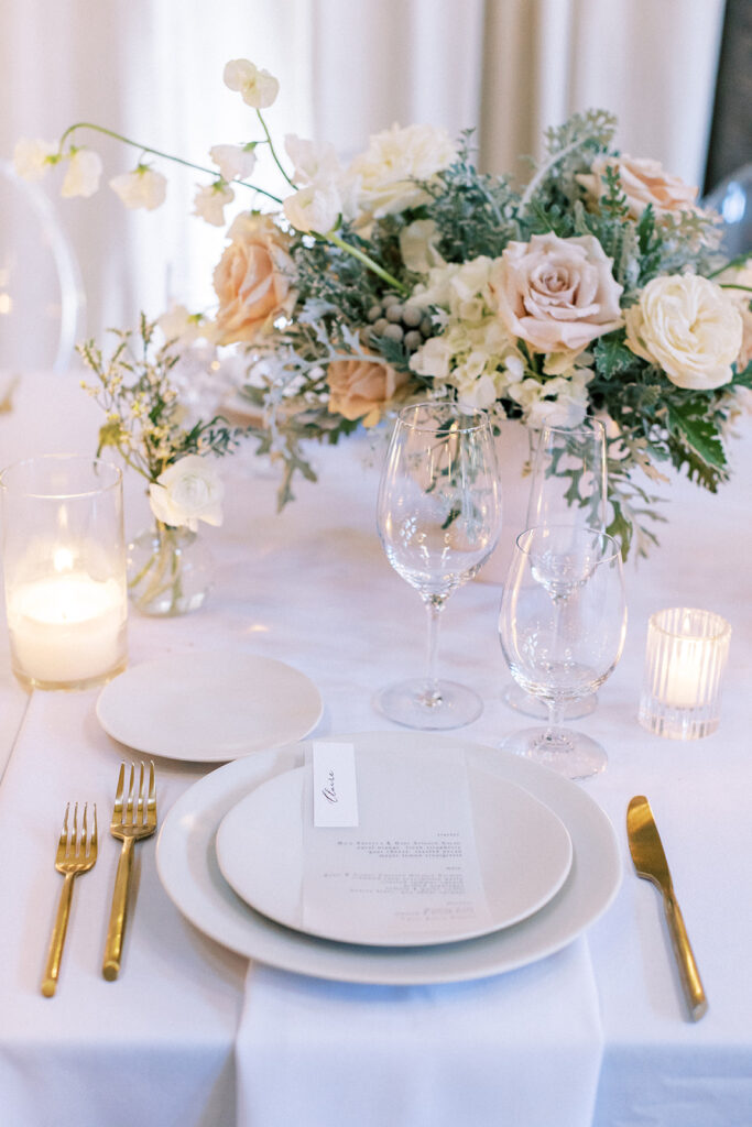 White and blush wedding design. Table setting with ceramic plates, gold flatware, and white linen tablecloth. Whimsical floral arrangement centerpiece with soft pink and white flowers.