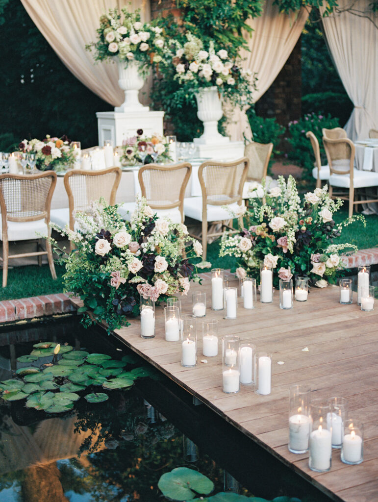 Wedding reception at the Belmond El Encanto hotel by the Lily Pond. Drapery hanging around the courtyard. White pillars with White urns full of white, pink, sand, and burgundy colored flowers. Candles lining the lily pond bridge.