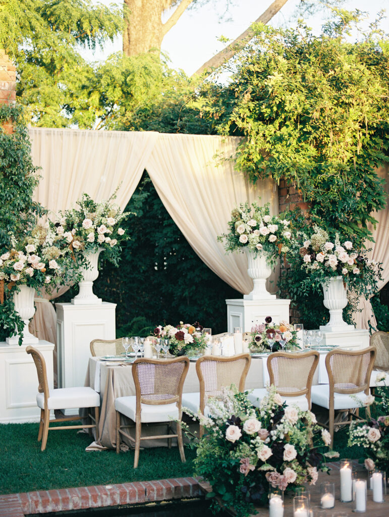 Wedding reception at the Belmond El Encanto hotel by the Lily Pond. Drapery hanging around the courtyard. White pillars with White urns full of white, pink, sand, and burgundy colored flowers. Candles lining the lily pond bridge. Green chargers and gold flatware.