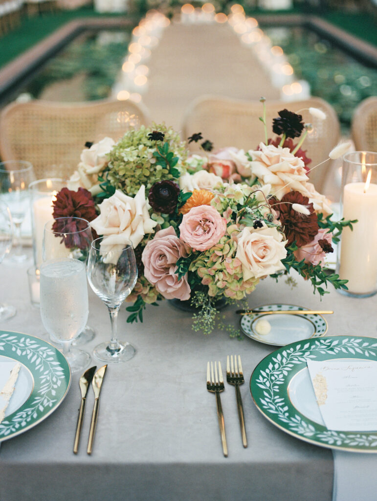 Wedding reception at the Belmond El Encanto hotel by the Lily Pond. Drapery hanging around the courtyard. White pillars with White urns full of white, pink, sand, and burgundy colored flowers. Candles lining the lily pond bridge. Green chargers and gold flatware.