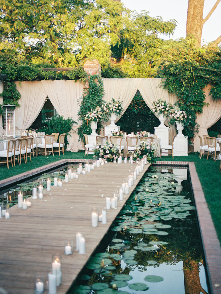 Wedding reception at the Belmond El Encanto hotel by the Lily Pond. Drapery hanging around the courtyard. White pillars with White urns full of white, pink, sand, and burgundy colored flowers. Candles lining the lily pond bridge.