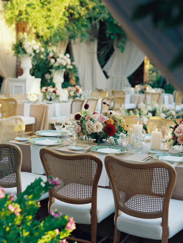 Wedding reception at the Belmond El Encanto hotel by the Lily Pond. Drapery hanging around the courtyard. White pillars with White urns full of white, pink, sand, and burgundy colored flowers. Candles lining the lily pond bridge. Green chargers and gold flatware.