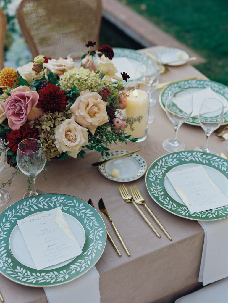 Wedding reception at the Belmond El Encanto hotel by the Lily Pond. Drapery hanging around the courtyard. White pillars with White urns full of white, pink, sand, and burgundy colored flowers. Candles lining the lily pond bridge. Green chargers, velvet linens and gold flatware.