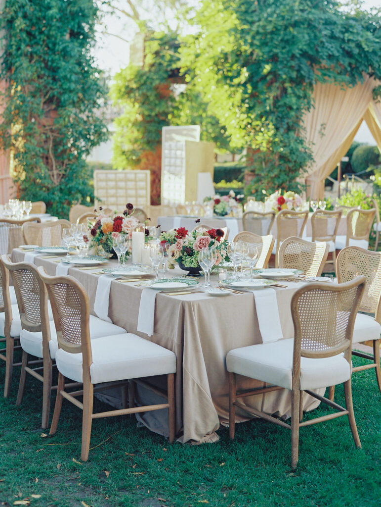 Wedding reception at the Belmond El Encanto hotel by the Lily Pond. Drapery hanging around the courtyard. White pillars with White urns full of white, pink, sand, and burgundy colored flowers. Candles lining the lily pond bridge. Green chargers, velvet linens and gold flatware.