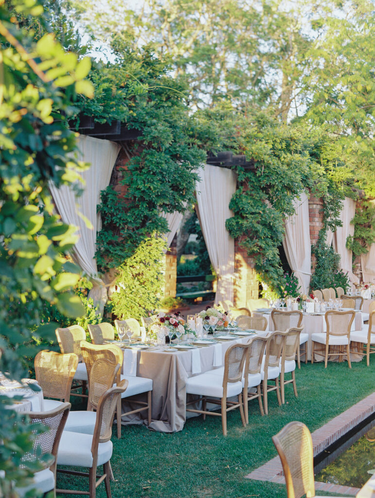 Wedding reception at the Belmond El Encanto hotel by the Lily Pond. Drapery hanging around the courtyard. White pillars with White urns full of white, pink, sand, and burgundy colored flowers. Candles lining the lily pond bridge. Green chargers, velvet linens and gold flatware.