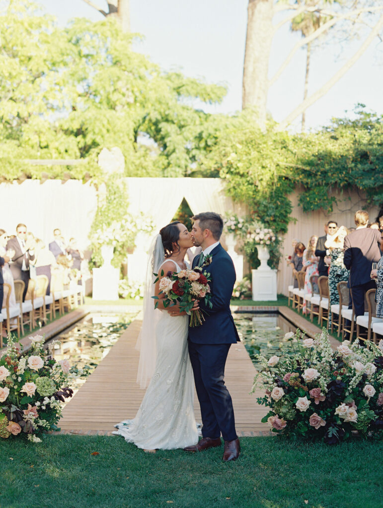 Wedding ceremony at the Belmond El Encanto hotel by the Lily Pond. Drapery hanging around the courtyard. White pillars with White urns full of white, pink, sand, and burgundy colored flowers.