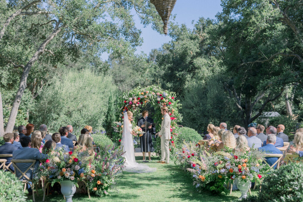 Home backyard garden wedding in Montecito with rainbow colored flowers on floral aisle, urns, and classic arch.