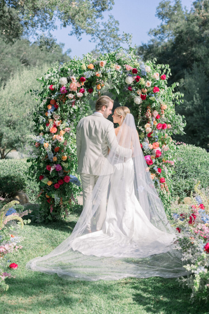 Home backyard garden wedding ceremony in Montecito with rainbow colored flowers on floral aisle, urns, and classic arch. Bride and Groom portrait.