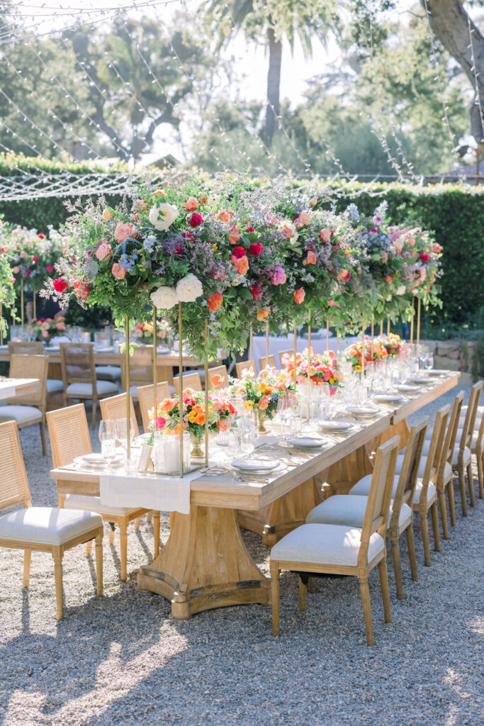 Montecito home Backyard wedding reception with lots of Colorful flowers and light wood furniture under a canopy of fairy lights