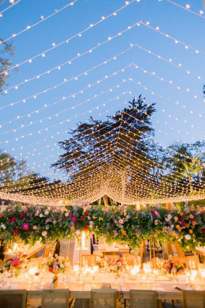 Montecito home Backyard wedding reception with lots of Colorful flowers and light wood furniture under a canopy of fairy lights