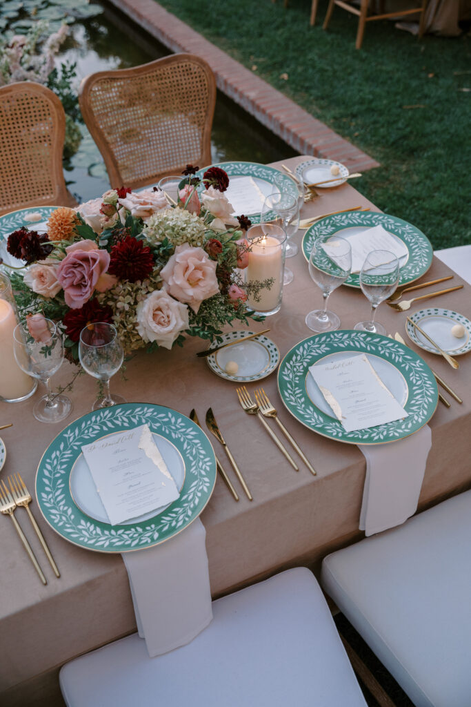 Wedding reception at the Belmond El Encanto hotel by the Lily Pond. Drapery hanging around the courtyard. White pillars with White urns full of white, pink, sand, and burgundy colored flowers. Candles lining the lily pond bridge. Green chargers, velvet linens and gold flatware.