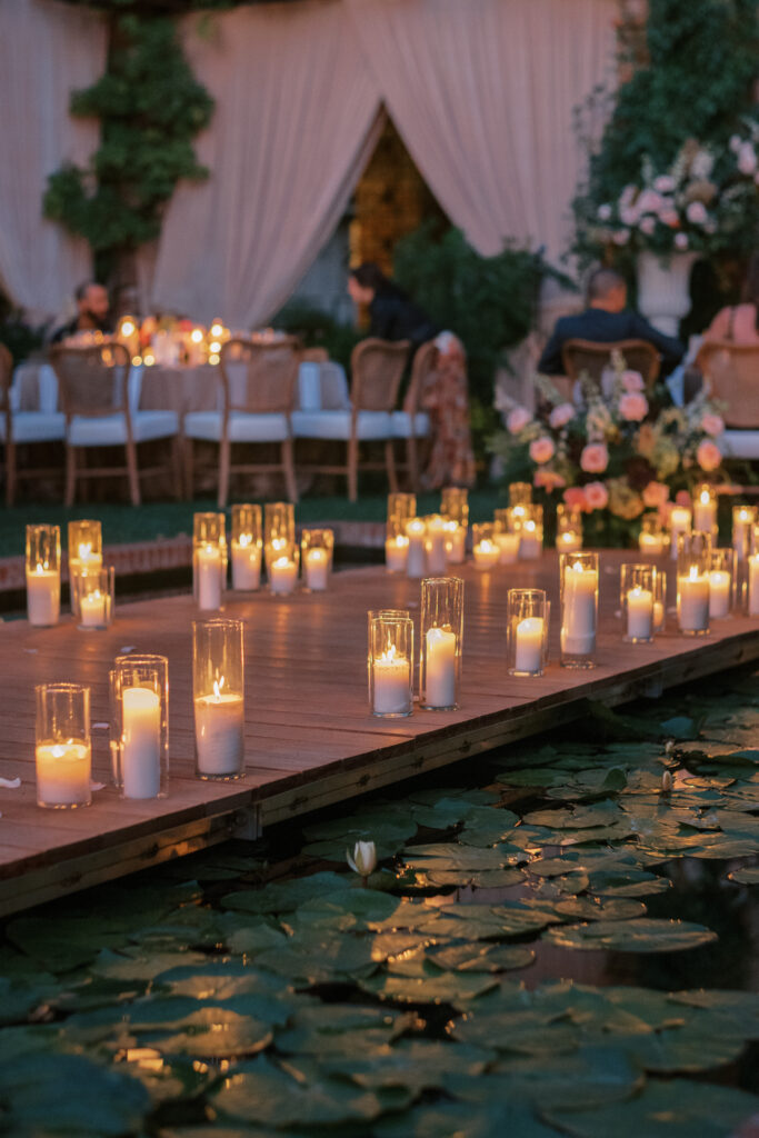 Wedding reception at the Belmond El Encanto hotel by the Lily Pond. Drapery hanging around the courtyard. White pillars with White urns full of white, pink, sand, and burgundy colored flowers. Candles lining the lily pond bridge. Green chargers, velvet linens and gold flatware.