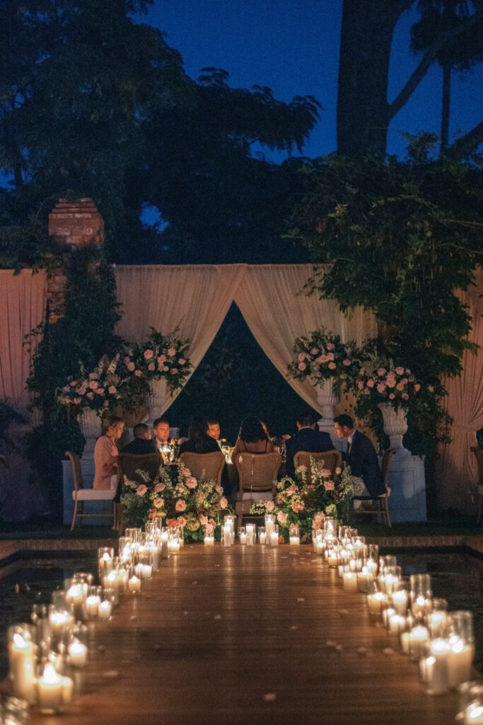 Wedding reception at the Belmond El Encanto hotel by the Lily Pond. Drapery hanging around the courtyard. White pillars with White urns full of white, pink, sand, and burgundy colored flowers. Candles lining the lily pond bridge. Green chargers, velvet linens and gold flatware.