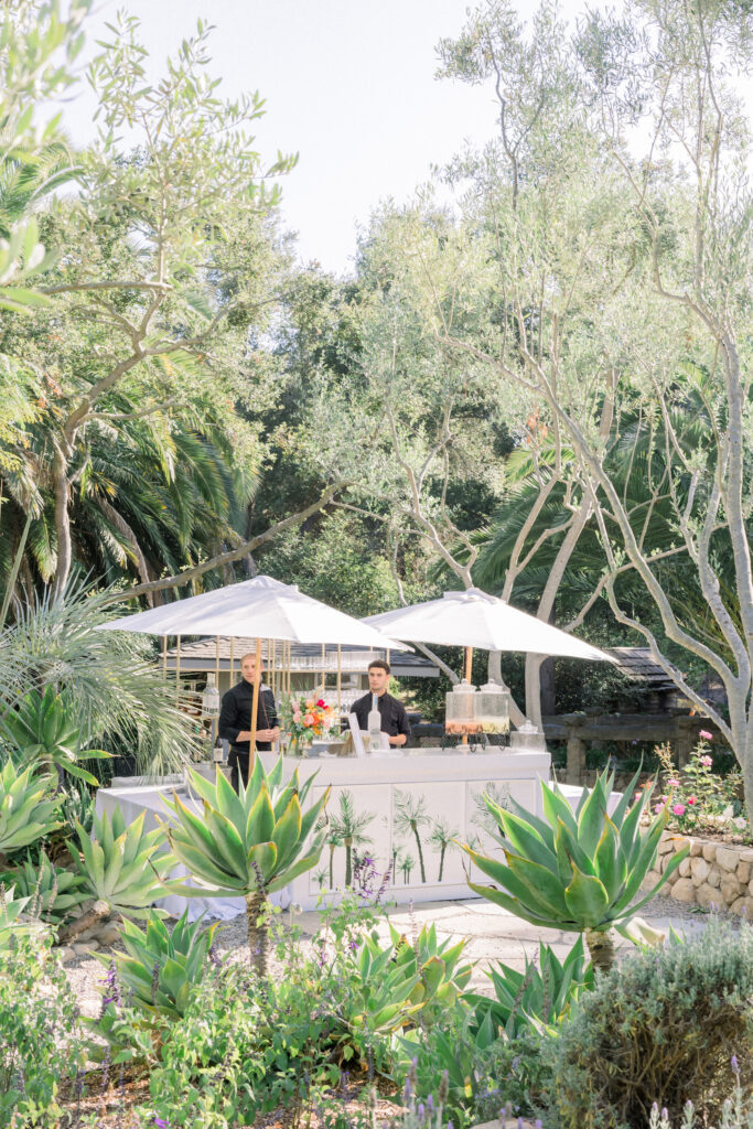 Home Backyard Wedding Cocktail hour in Montecito. White outdoor rental bar with palm trees painted on front.