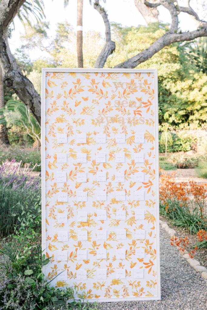 Custom wedding seating chart wall display escort cards. Framed white wall with painted orange and gold floral pattern.