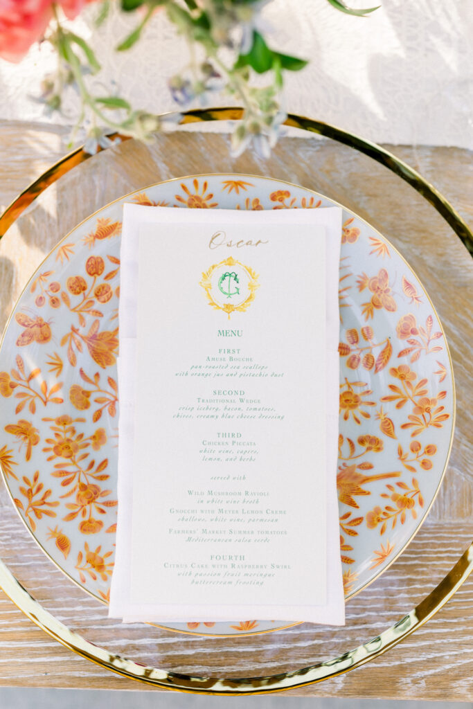 Wedding reception with Colorful flowers and light wood table. Orange patterned dinner plate on gold rimmed charger with custom monogrammed menu.