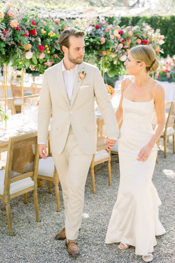 Bride and Groom walking their Montecito home Backyard wedding reception with lots of Colorful flowers and light wood furniture under a canopy of fairy lights