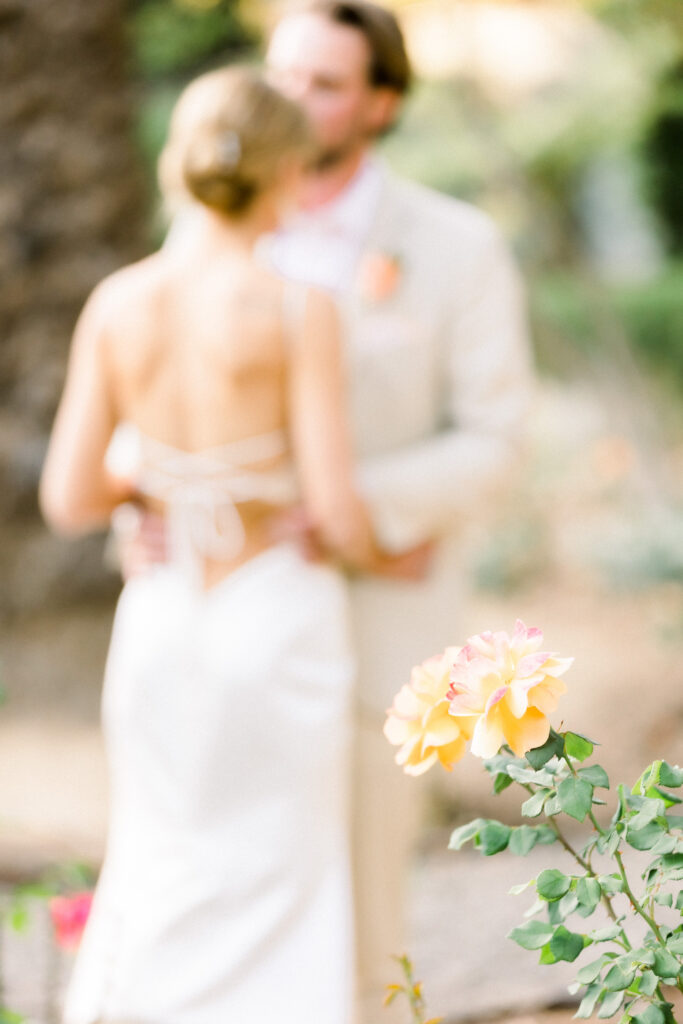 Bride and Groom portrait in Montecito home backyard garden at sunset. Groom in light beige suit and brown dress shoes. Bride with low bun and criss-cross back.