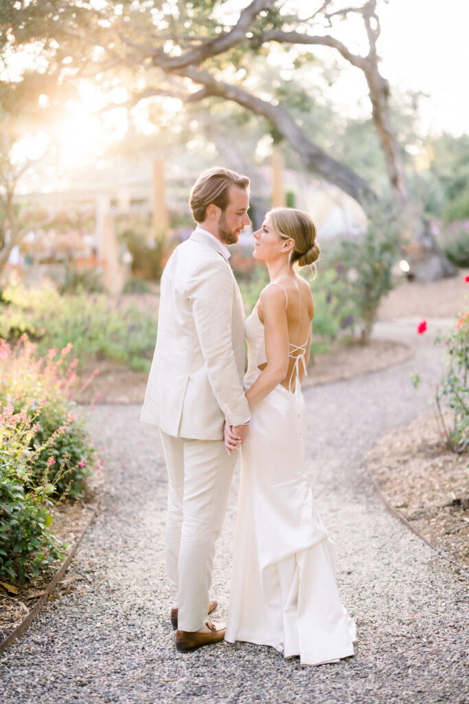 Bride and Groom portrait in Montecito home backyard garden at sunset. Groom in light beige suit and brown dress shoes. Bride with low bun and criss-cross back.