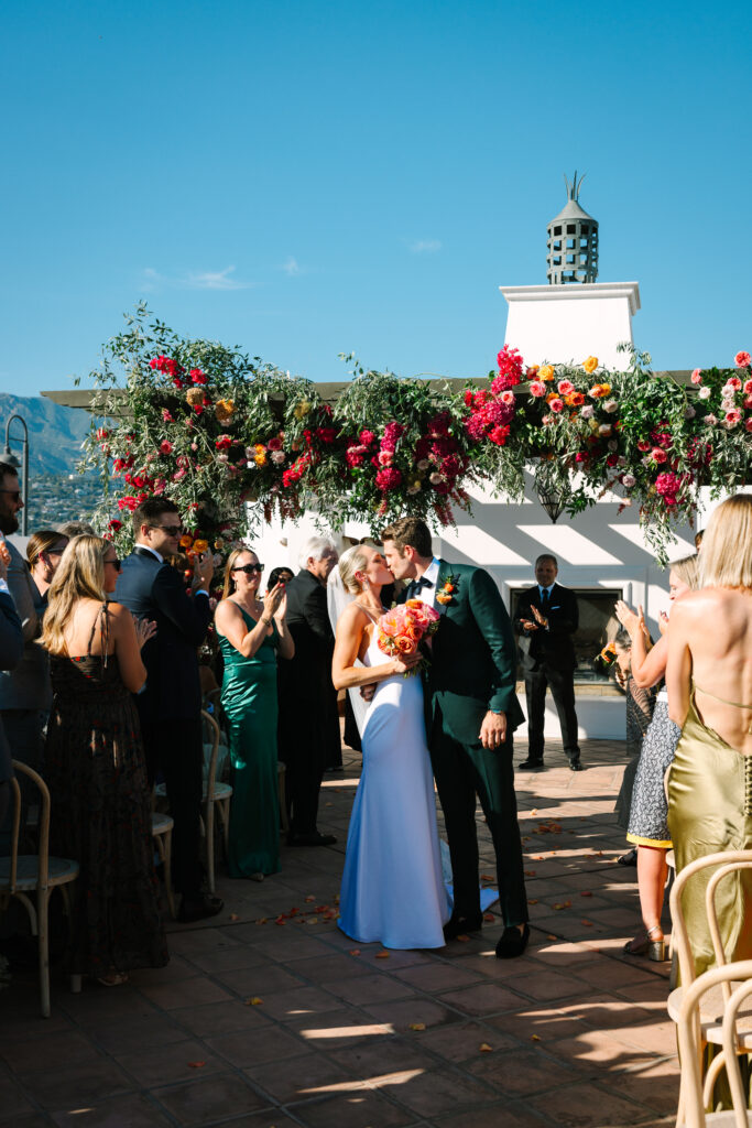 Wedding ceremony on Hotel Californian rooftop with large pink and orange floral arch. Bride and groom kissing down the aisle