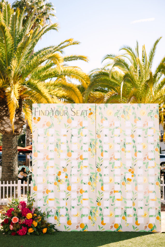 Wedding seating chart. Orange patterned escort card display wall with pink flowers in front and palm trees in background. Outside at Hotel Californian.