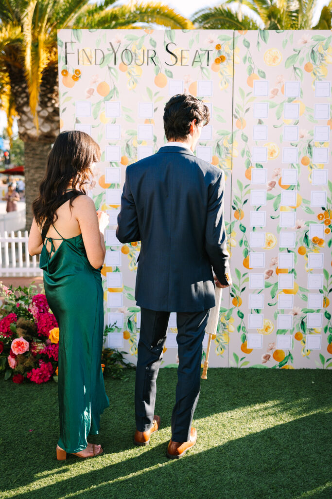 Wedding seating chart. Orange patterned escort card display wall with pink flowers in front and palm trees in background. Outside at Hotel Californian.