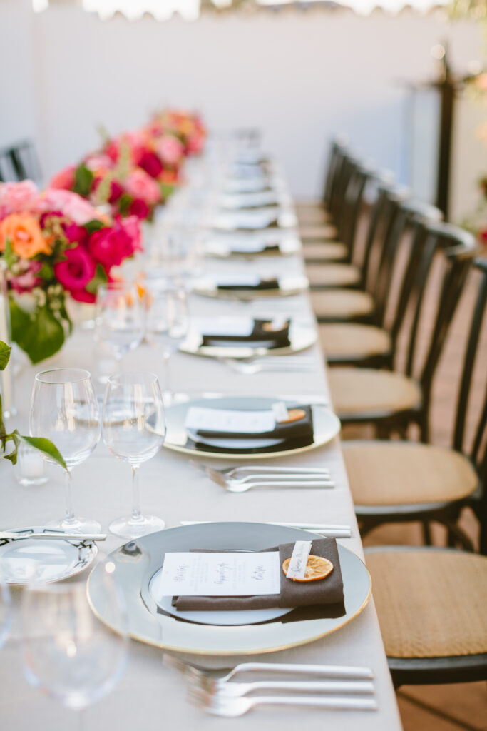 Wedding table scape with white table linen, black chargers and napkins with dried oranges, pink and orange floral arrangements as centerpieces.