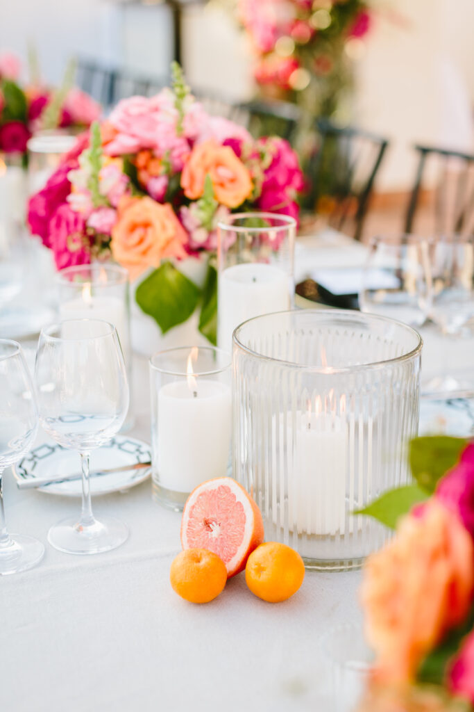 Wedding table scape with white table linen, black chargers and napkins, pink and orange floral arrangements as centerpieces with cut citrus around candles, and a floral installation in the background.