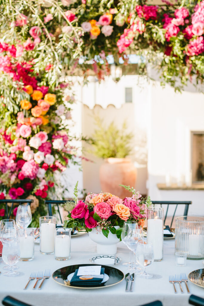 Wedding table scape with white table linen, black chargers and napkins, pink and orange floral arrangements as centerpieces, and a floral installation in the background.