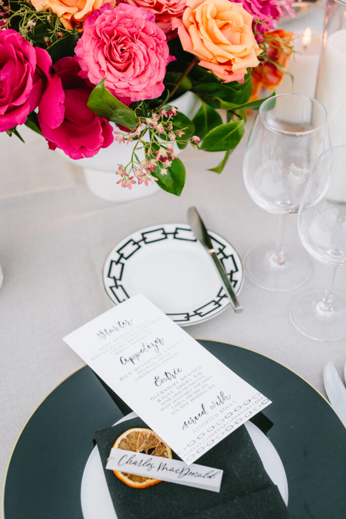 Wedding table scape with white table linen, black chargers and napkins, pink and orange floral arrangements as centerpieces, and a floral installation in the background. Menu design that matches the dinner plates.