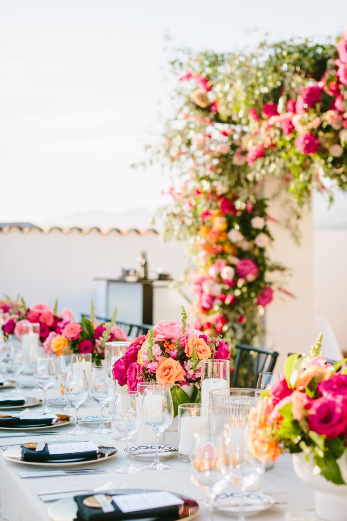 Wedding table scape with white table linen, black chargers and napkins, pink and orange floral arrangements as centerpieces, and a floral installation in the background. Wedding tablescape.