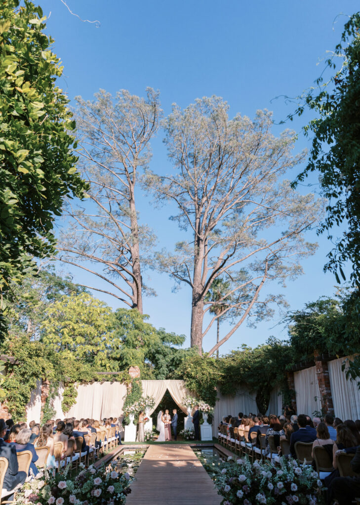 Wedding ceremony at the Belmond El Encanto hotel by the Lily Pond. Drapery hanging around the courtyard. White pillars with White urns full of white, pink, sand, and burgundy colored flowers.