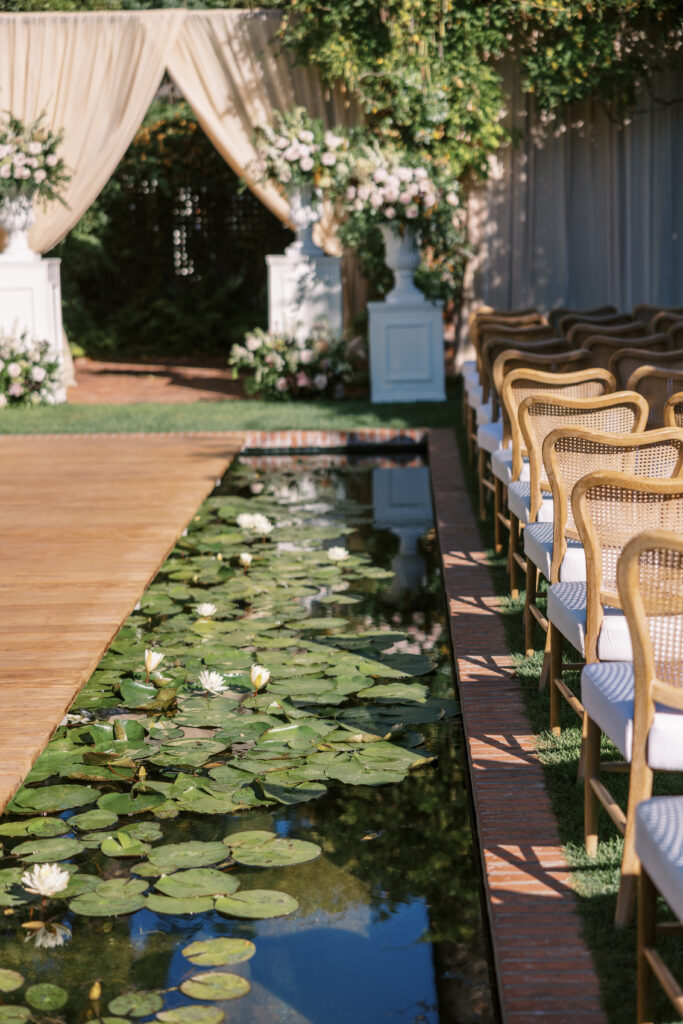 Wedding ceremony at the Belmond El Encanto hotel by the Lily Pond. Drapery hanging around the courtyard. White pillars with White urns full of white, pink, sand, and burgundy colored flowers.