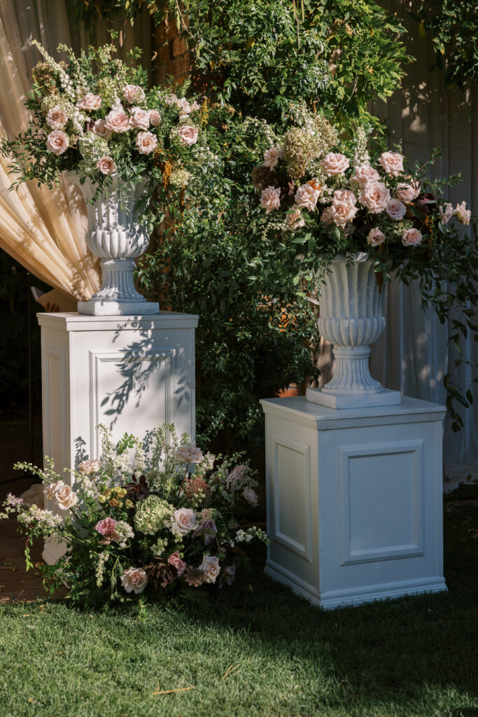 Wedding ceremony at the Belmond El Encanto hotel by the Lily Pond. Drapery hanging around the courtyard. White pillars with White urns full of white, pink, sand, and burgundy colored flowers.