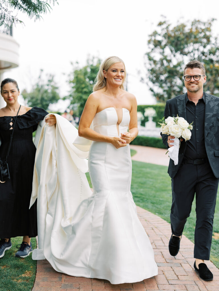 Tyler Speier and team member holding bouquet and helping Bride walk in her wedding dress at the Rosewood Miramar Beach resort