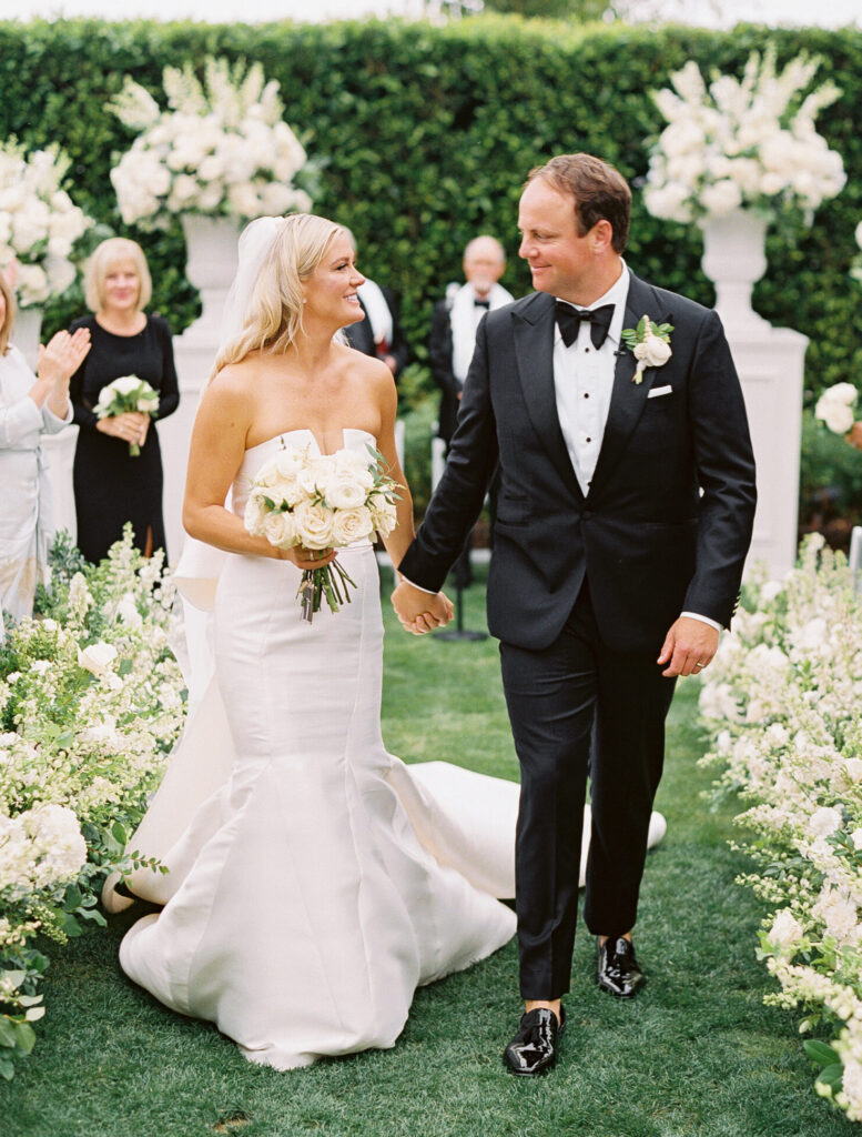 Portrait of the Bride and Groom walking down white floral aisle after wedding ceremony at the Rosewood Miramar Beach Resort