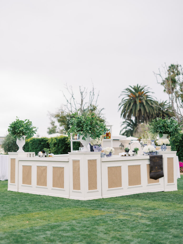 Large custom bar on the Rosewood Miramar Beach lawn at wedding cocktail hour with urns full of greenery and blue and white patterned vases