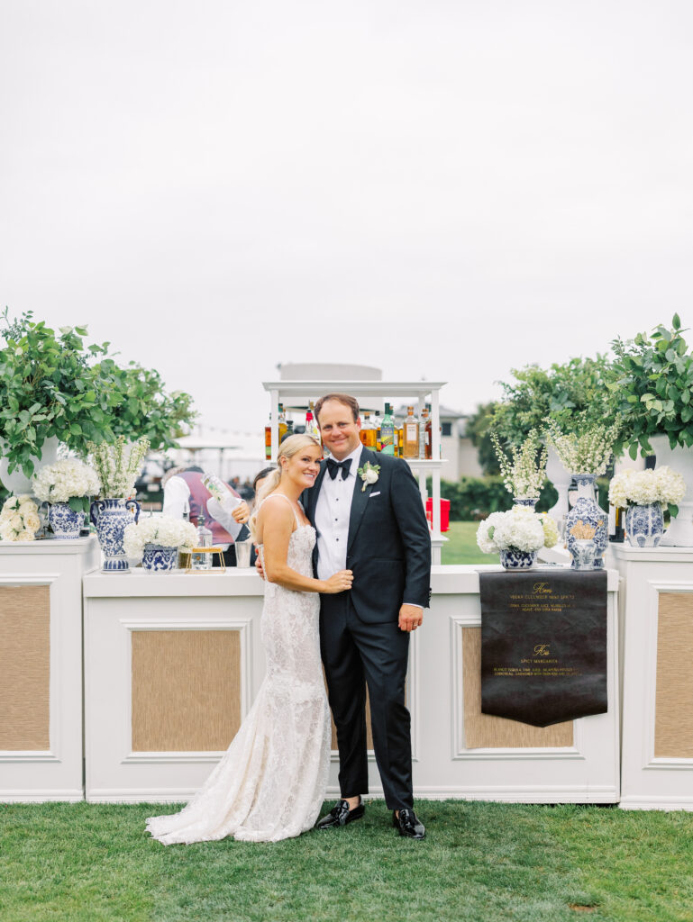 Bride and groom in front of large custom bar on the Rosewood Miramar Beach lawn at wedding cocktail hour with urns full of greenery and blue and white patterned vases