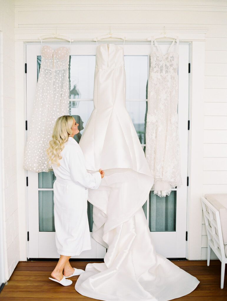 Bride in robe and slippers getting ready with her three bridal gowns hanging up on door at Rosewood Miramar Beach Resort