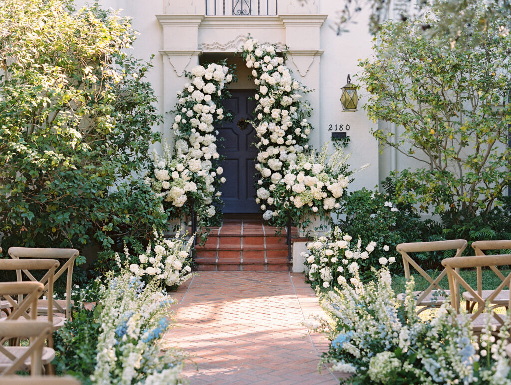 At home wedding ceremony in front of house. Deconstructed ceremony arch and curvy floral aisle with white and blue flowers.