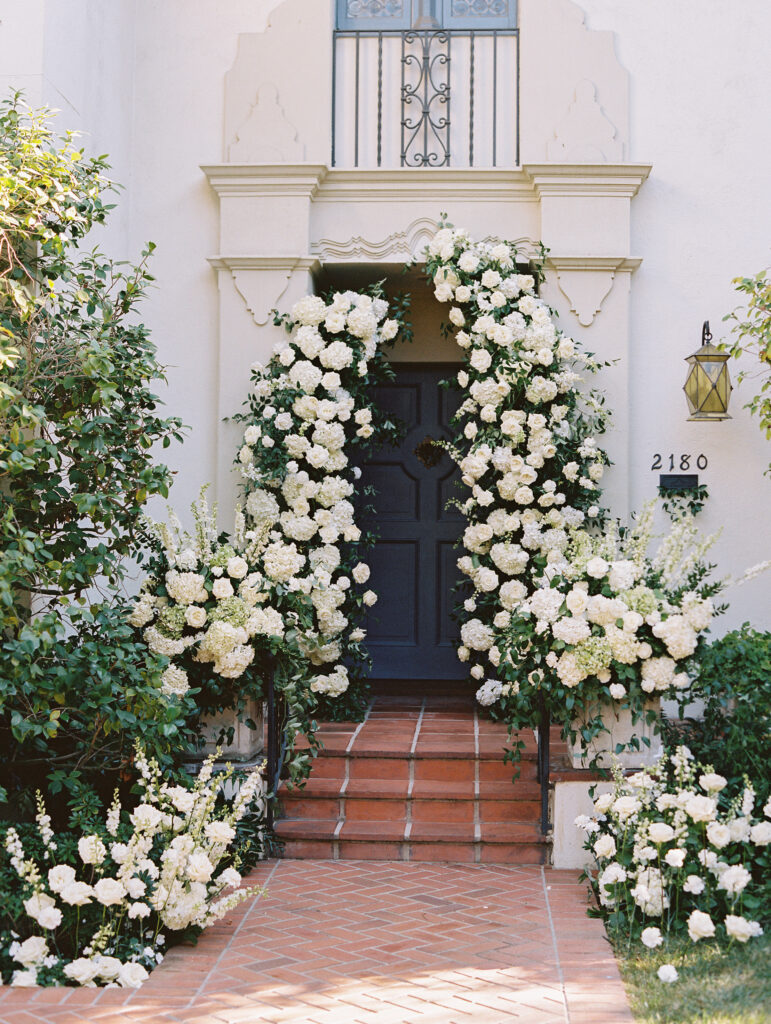 At home wedding ceremony in front of house. Deconstructed ceremony arch and curvy floral aisle with white and blue flowers.