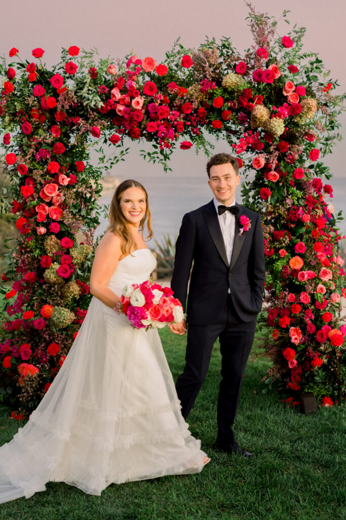 Vibrant pink and red wedding arch with roses and bougainvillea. Bride and groom walking in front of the arch with bright pink bridal bouquet and boutonnière.