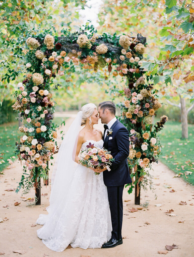 Fall wedding ceremony at Kestrel Park in Santa Ynez. Very tall wedding arch covered in fall colored flowers and foliage in front of an allay of fall colored trees.