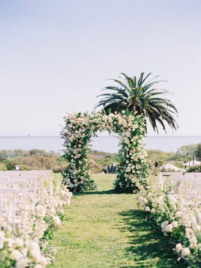 Wedding ceremony at The Ritz Carlton Bacara with ocean view in background. Arch and floral aisle with lush white flowers.