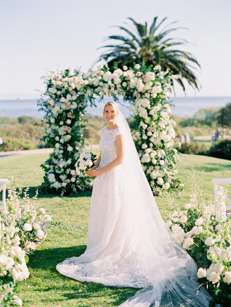 Wedding ceremony at The Ritz Carlton Bacara with ocean view in background. Arch and floral aisle with lush white flowers. Bride standing in aisle.