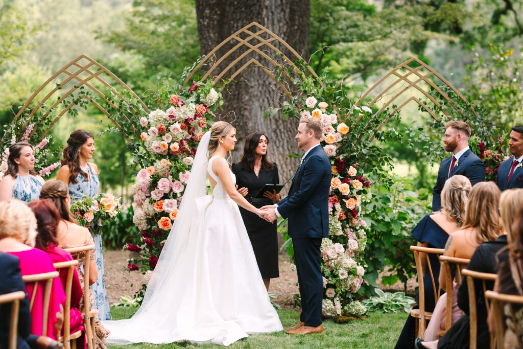 Wedding ceremony at Beaulieu Garden, Napa. Custom triple arch with burgundy and pink flowers. Bride and groom saying vows.