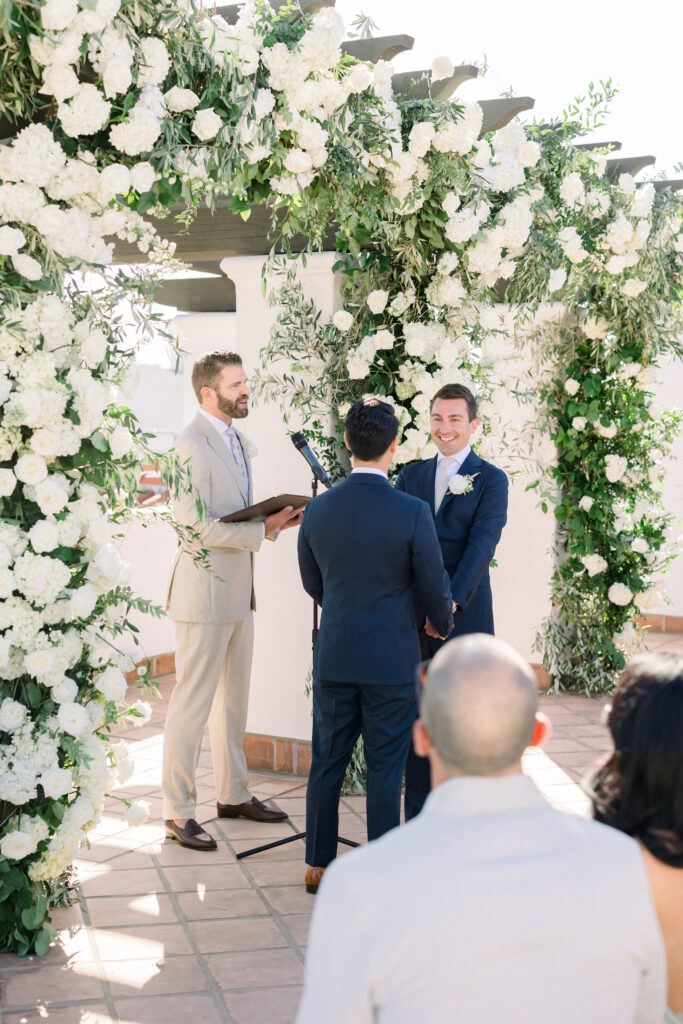 Wedding ceremony at Hotel Californian with triple arch with white flowers. Two grooms in blue suits saying wedding vows under arch.