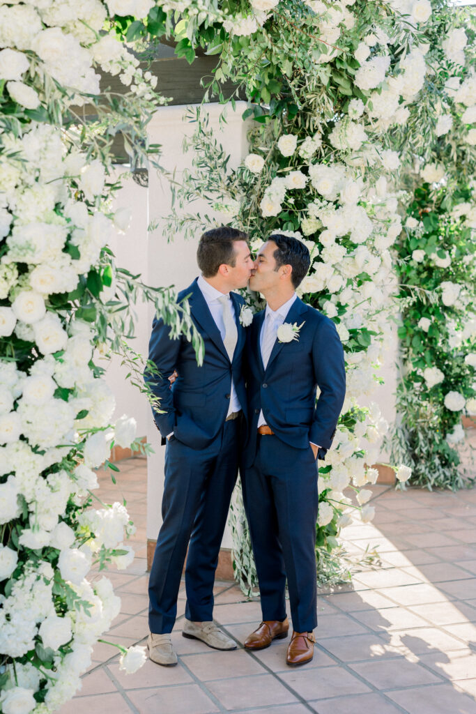 Wedding ceremony at Hotel Californian with triple arch with white flowers. Two grooms in blue suits saying wedding vows under arch.