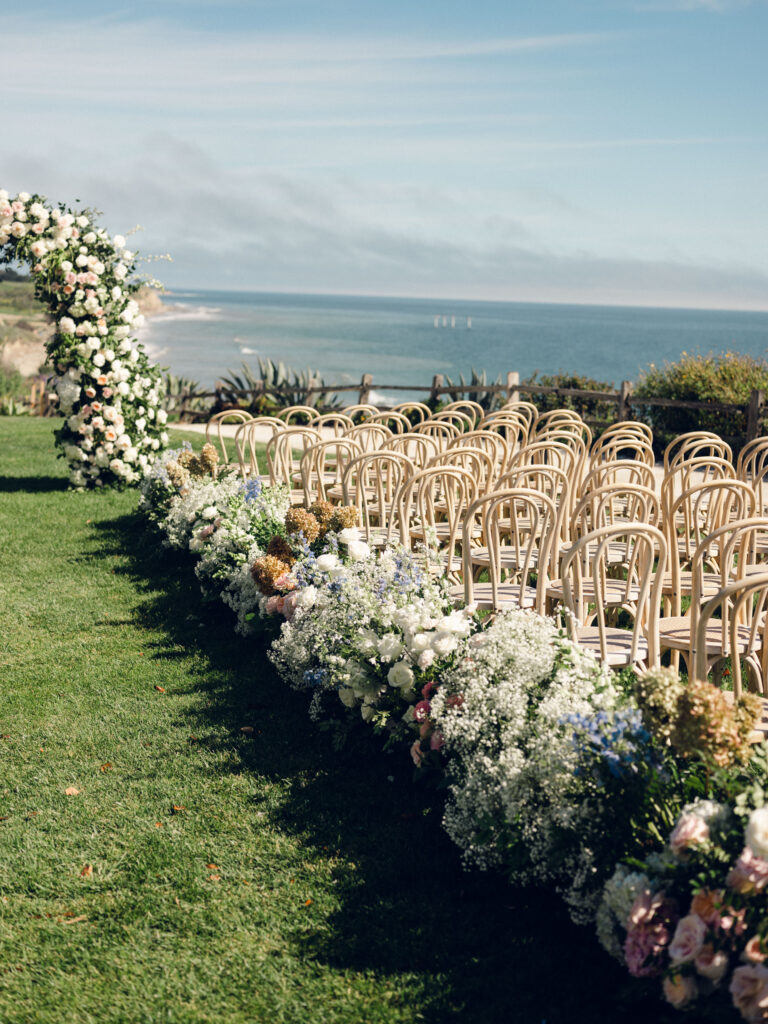 Wedding ceremony at The Ritz Carlton Bacara with ocean view in background. Large curved wedding arch with fluffy white and pink flowers. Light wood ceremony chairs. Fluffy full floral aisle with baby's breath, hydrangea, and roses.