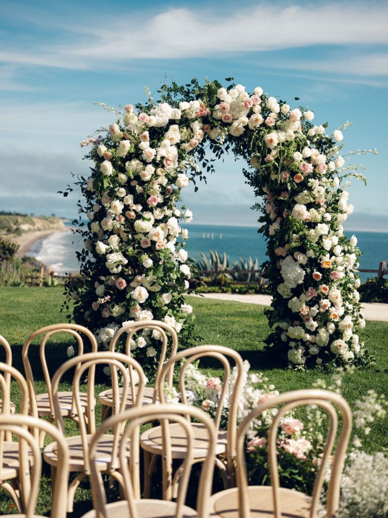 Wedding ceremony at The Ritz Carlton Bacara with ocean view in background. Large curved wedding arch with fluffy white and pink flowers. Light wood ceremony chairs.